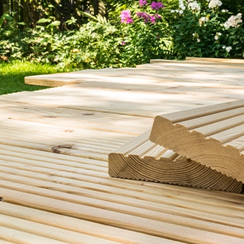 Terrassenbau mit hellen Lärchenholzdielen, gestapelte Riffelbretter im Vordergrund, umgeben von grünem Garten.