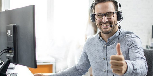 Freundlicher Kundenberater mit Headset sitzt an einem Schreibtisch mit Computer und zeigt den Daumen hoch.