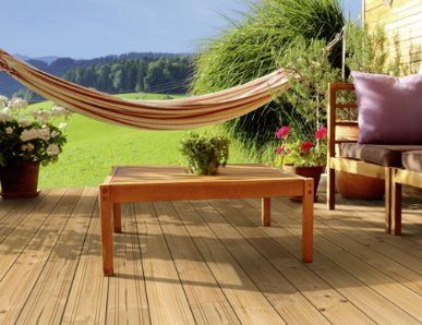 Gemütliche Holzterrasse mit Douglasien-Dielen, Lounge-Sofa mit Kissen, Holztisch, Hängematte und blühenden Pflanzen vor idyllischer Landschaft.