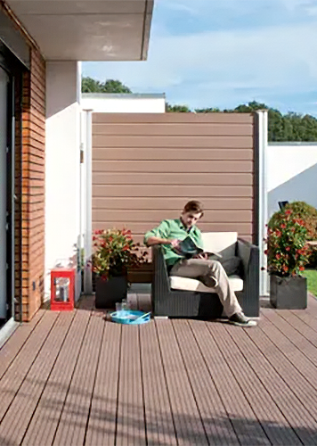 Eine Person sitzt in einem Gartensessel auf einer Holzterrasse und liest Zeitung.